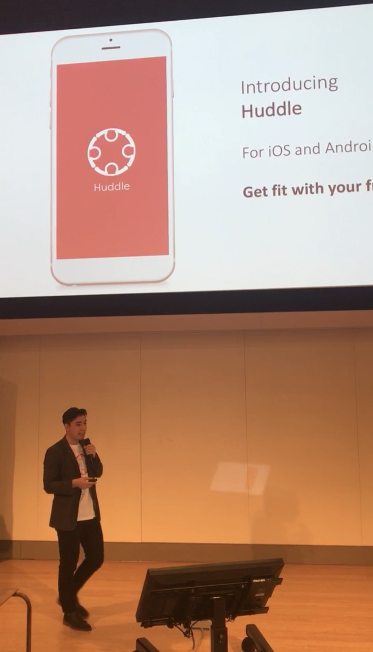 Dylan on stage with a microphone, presenting a slide that reads “Introducing Huddle — for iOS and Android”.
